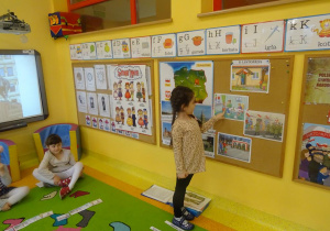 Dziewczynka stoi pod tablicą i wskazuje jedną z ilustracji, opowiada co na niej widać.